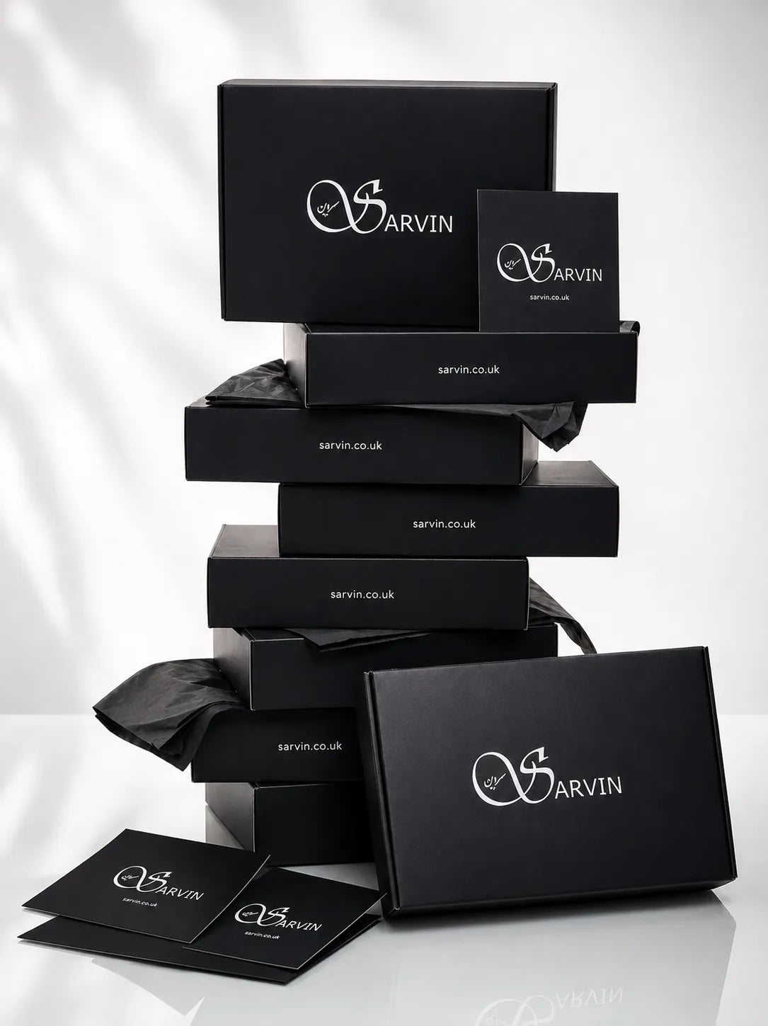 A selection of Sarvin branded black gift boxes in various sizes is neatly arranged on a flat surface. Positioned in front are two Sarvin Gift Cards featuring elegant white lettering, ideal for anyone looking for gift cards UK. The scene is set against a softly lit, neutral background.