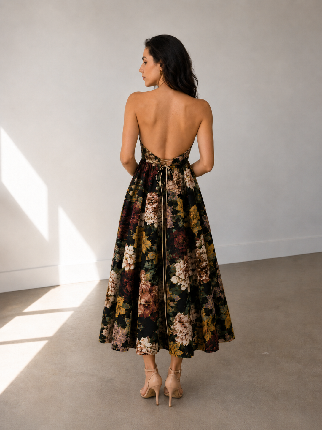 A woman stands indoors on a bare floor, facing away from the camera, wearing the Sarvin Golshifteh Floral Corset Midi Dress and nude high heels. The background and lighting are neutral and minimal.