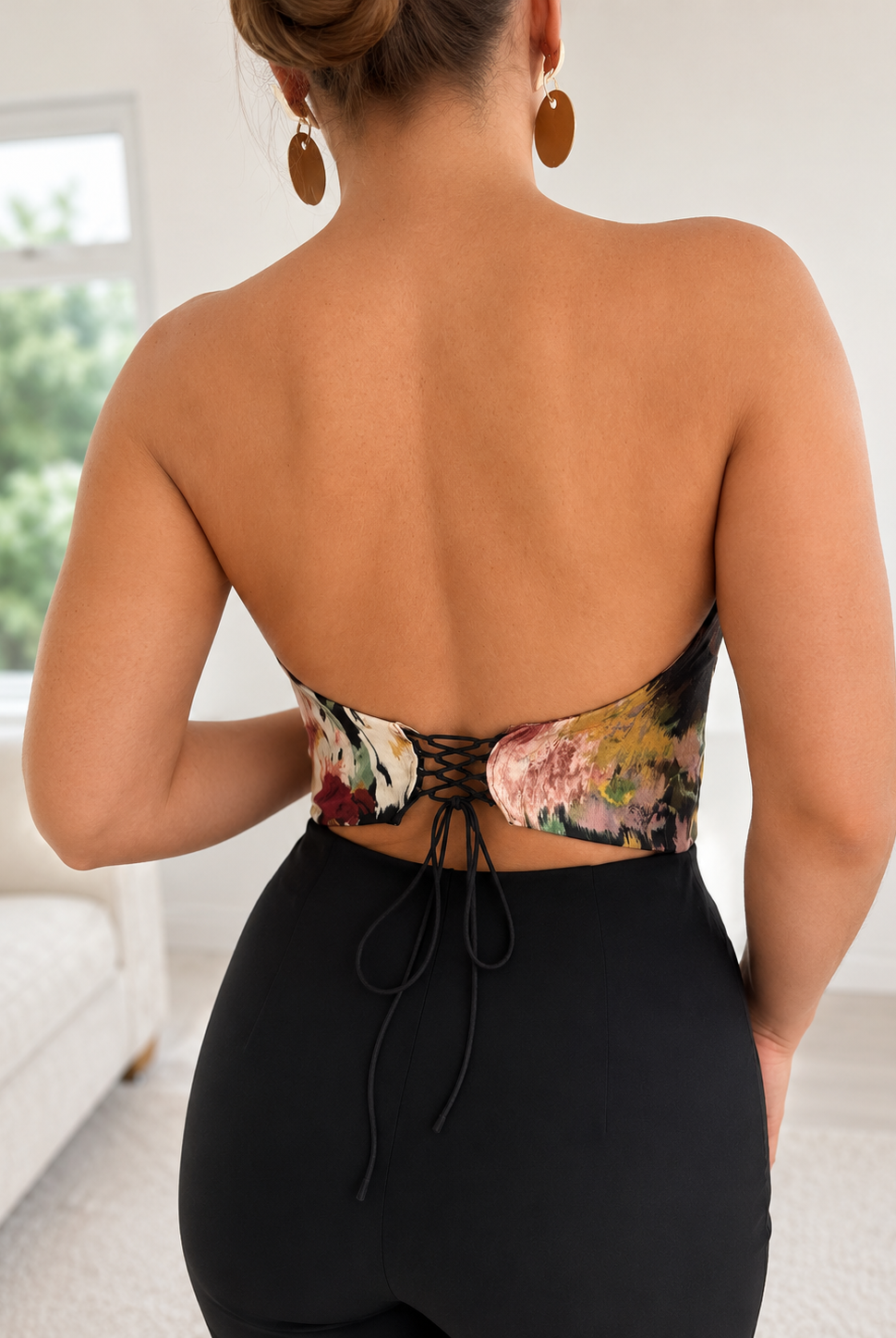 A person stands indoors with their back to the camera, wearing the Sarvin Ivy Corset Top—a waist-cinching floral halter style with lace-up detail—paired with high-waisted black pants. Their hair is up and they wear large round earrings.