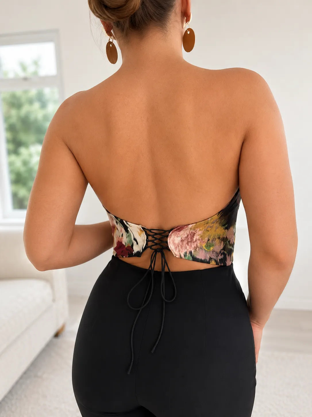 A person stands indoors with their back to the camera, wearing the Sarvin Ivy Corset Top—a waist-cinching floral halter style with lace-up detail—paired with high-waisted black pants. Their hair is up and they wear large round earrings.