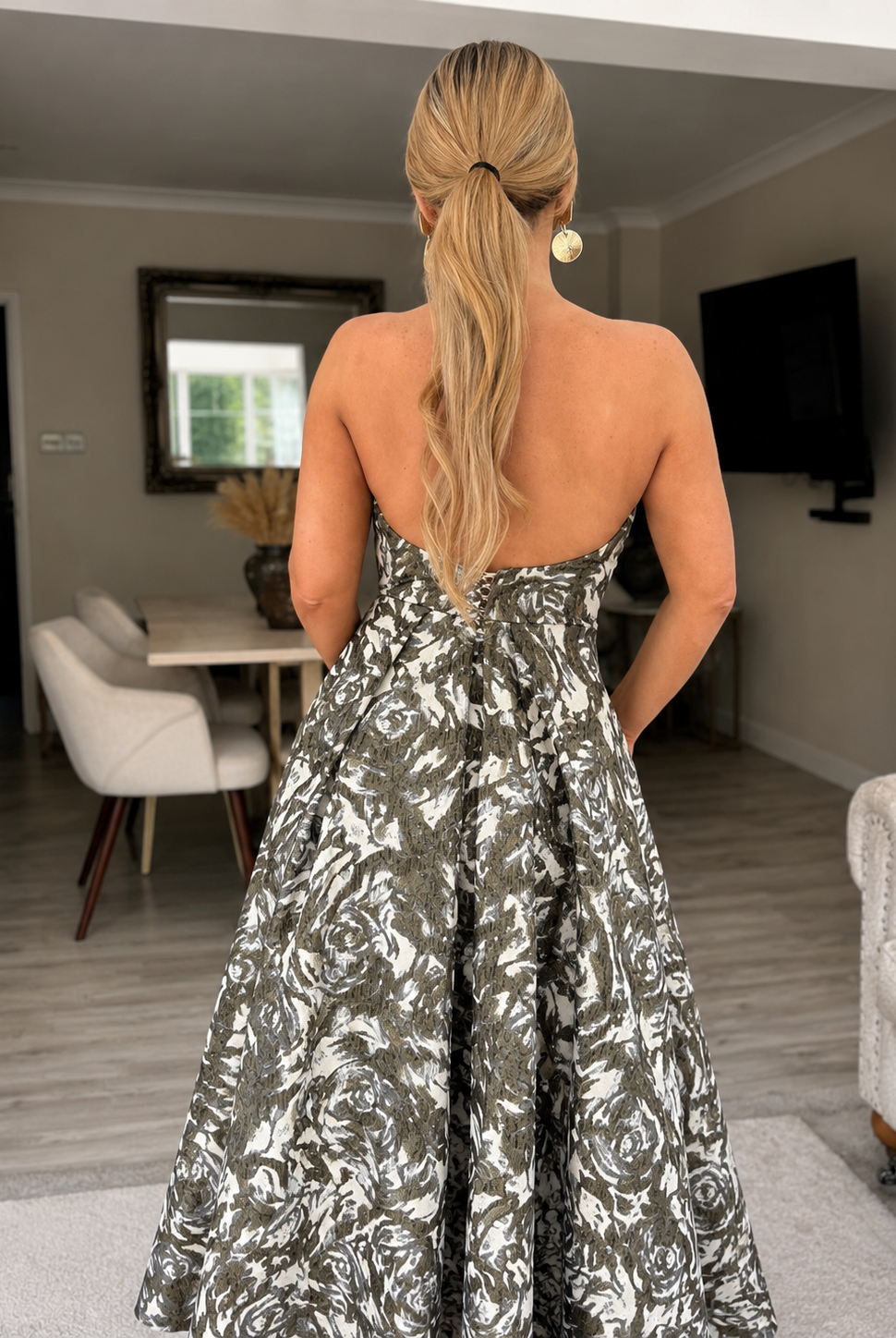 A woman with long blonde hair in a low ponytail stands indoors, facing away, wearing the Sarvin Tala Corset Midi Dress—a strapless black, white, and gray floral piece—in a modern living and dining area.