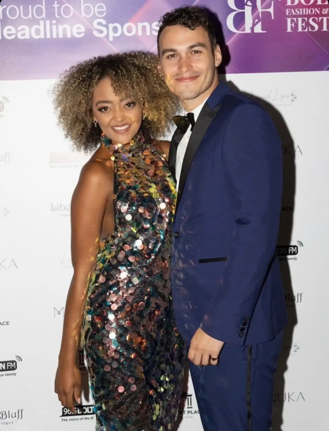 A woman wearing the Sarvin High Neck Backless Sequin Mini Dress poses and smiles next to a man in a blue suit and bow tie, both standing in front of an event step-and-repeat backdrop.