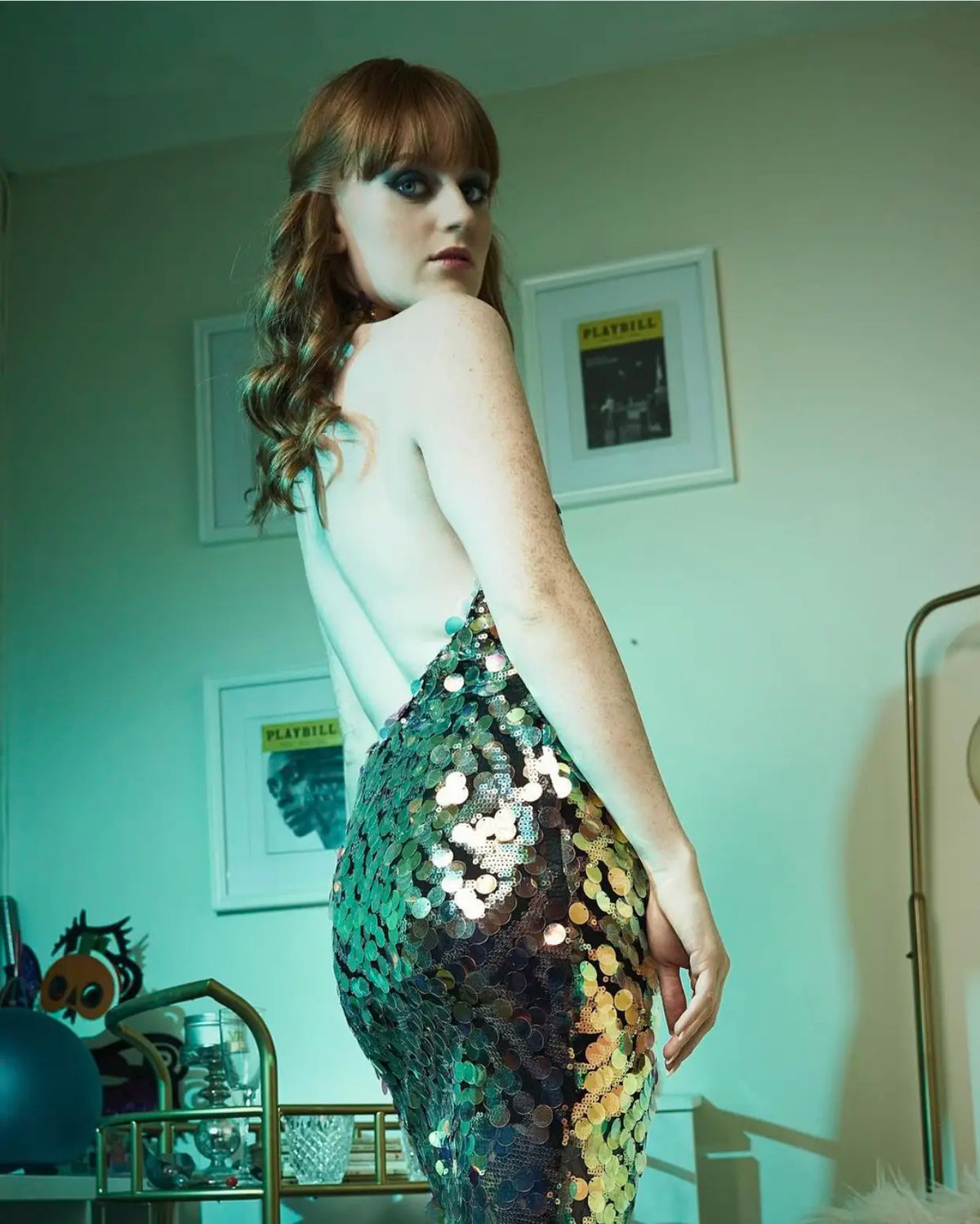 A person with long red hair stands in a room, wearing the Sarvin High Neck Backless Sequin Mini Dress. Framed Playbill posters are on the wall behind them, illuminated by greenish-blue lighting.
