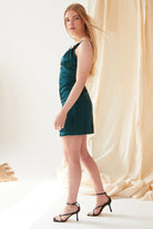 A woman with long blonde hair poses sideways in a Sarvin Emerald Green Mini Dress and black high-heeled sandals, standing before a light beige fabric backdrop in a well-lit room.