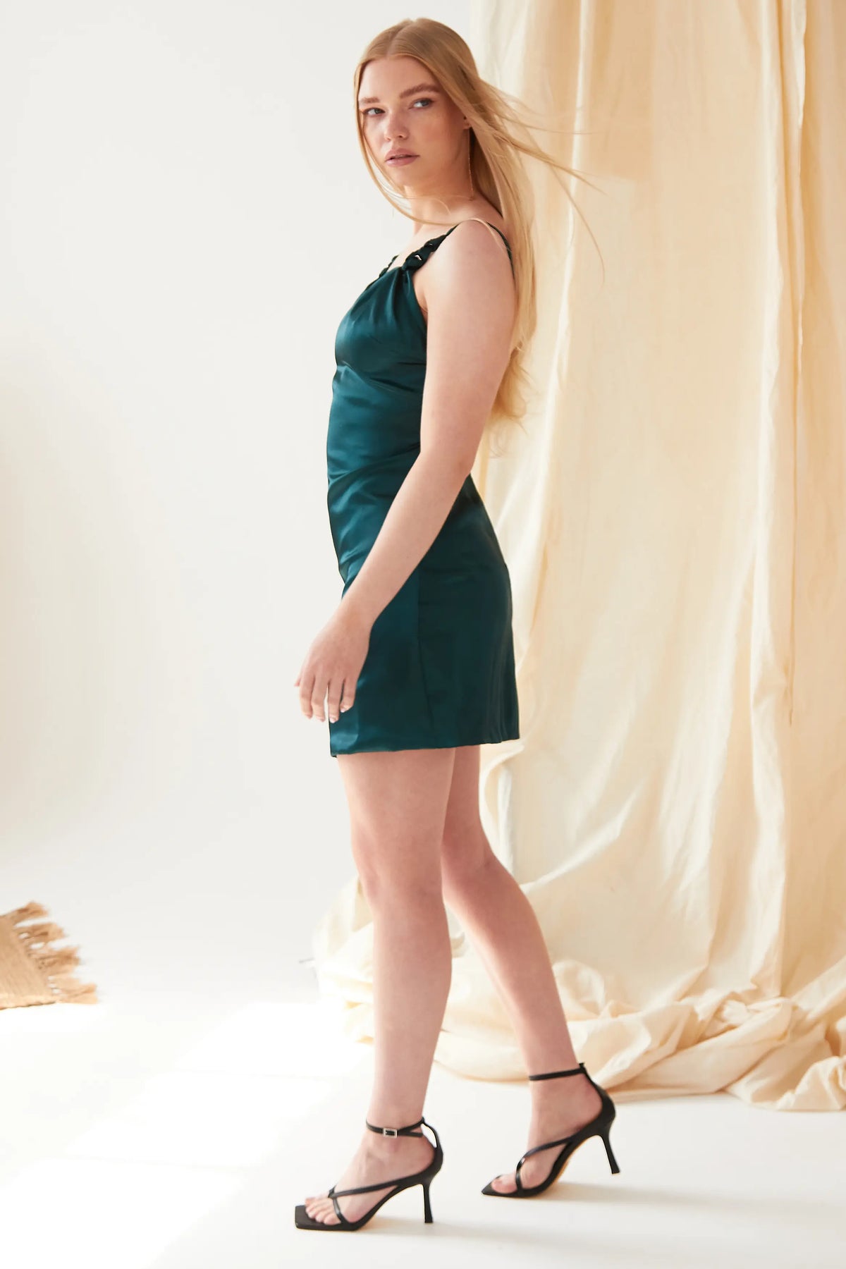 A woman with long blonde hair stands in profile wearing the Sarvin Emerald Green Silk Satin Backless Mini Dress and black strappy heels, indoors before a beige fabric backdrop in soft lighting.