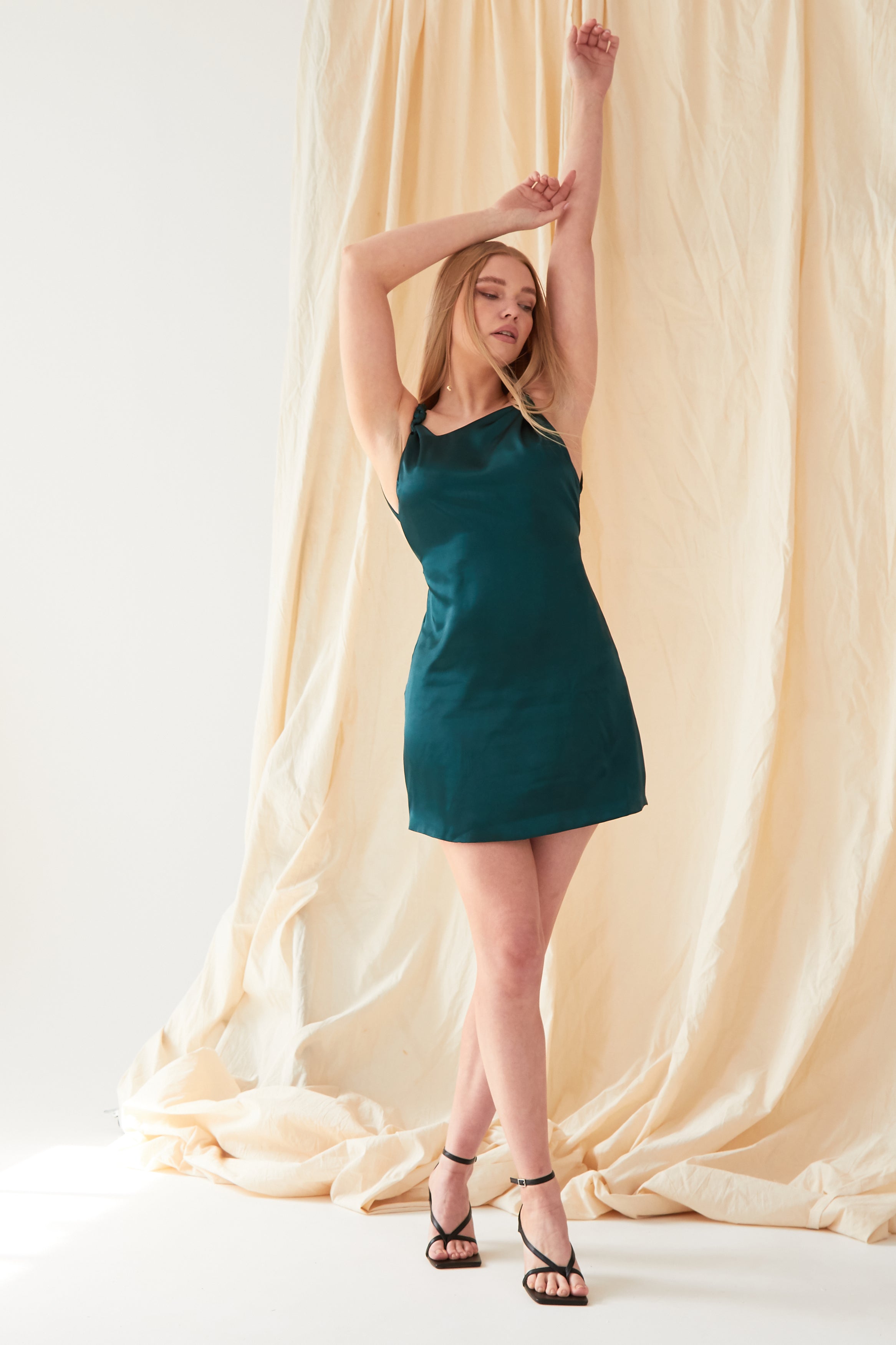 A woman wearing the Sarvin Emerald Green Mini Dress stands before a beige draped backdrop, arms raised above her head.
