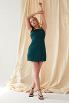 A woman wears the Sarvin Emerald Green Mini Dress, standing before a beige fabric backdrop with arms raised and one leg forward, paired with black strappy sandals.