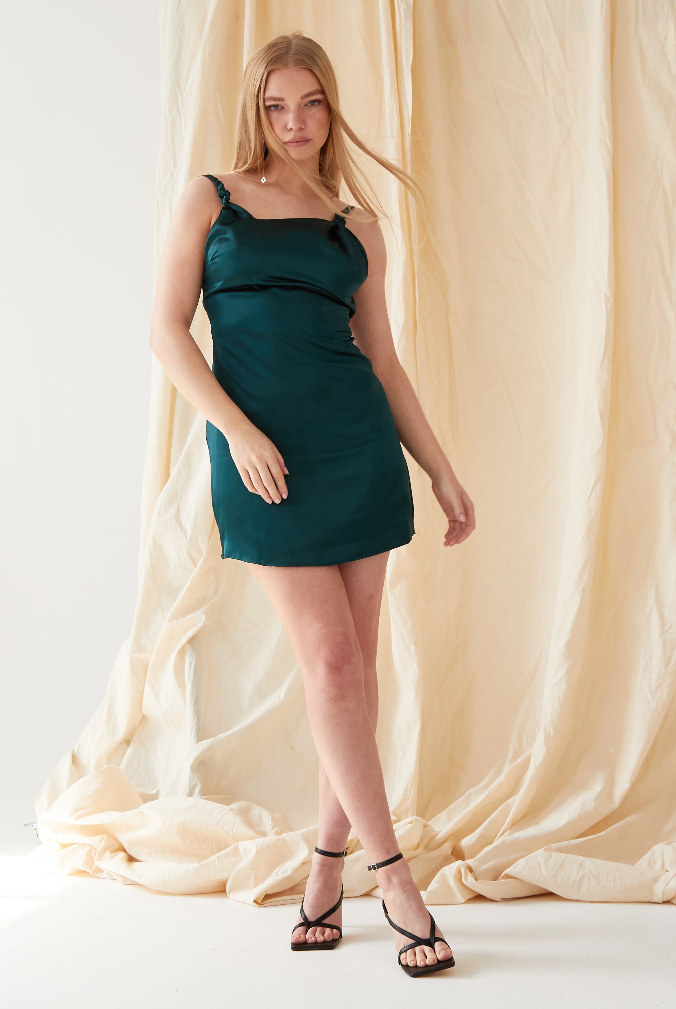 A woman with long blonde hair models the Sarvin Emerald Green Mini Dress, paired with black heeled sandals. She poses in front of a beige draped fabric backdrop, one arm relaxed and the other slightly bent.