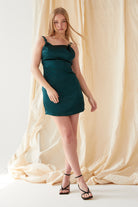 A woman with long blonde hair models the Sarvin Emerald Green Mini Dress, paired with black heeled sandals. She poses in front of a beige draped fabric backdrop, one arm relaxed and the other slightly bent.