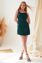 A woman with long blonde hair models the Sarvin Emerald Green Mini Dress, paired with black strappy heels. She stands indoors with one leg crossed, surrounded by neutral decor featuring pampas grass and draped fabric.
