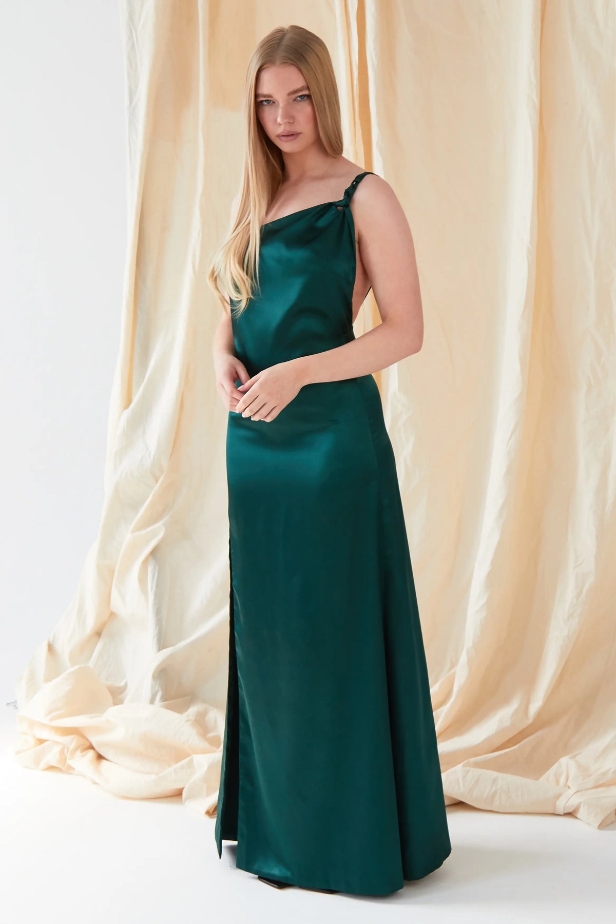 A woman with long straight blonde hair stands before beige draped fabric, wearing the Sarvin Emerald Green Maxi Dress in satin, featuring thin straps and a side slit.