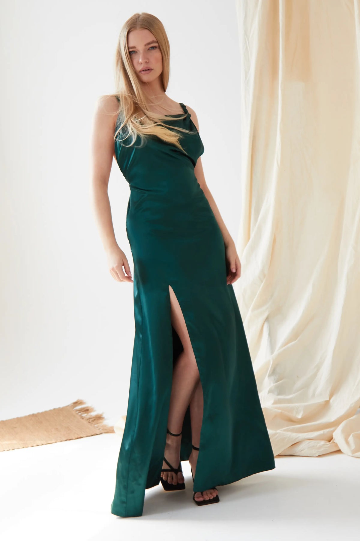 A woman wears the Sarvin Emerald Green Maxi Dress, a sleeveless satin gown with a thigh-high slit. She pairs it with black heels as her long blonde hair flows and delicate fabric drapes in the background.