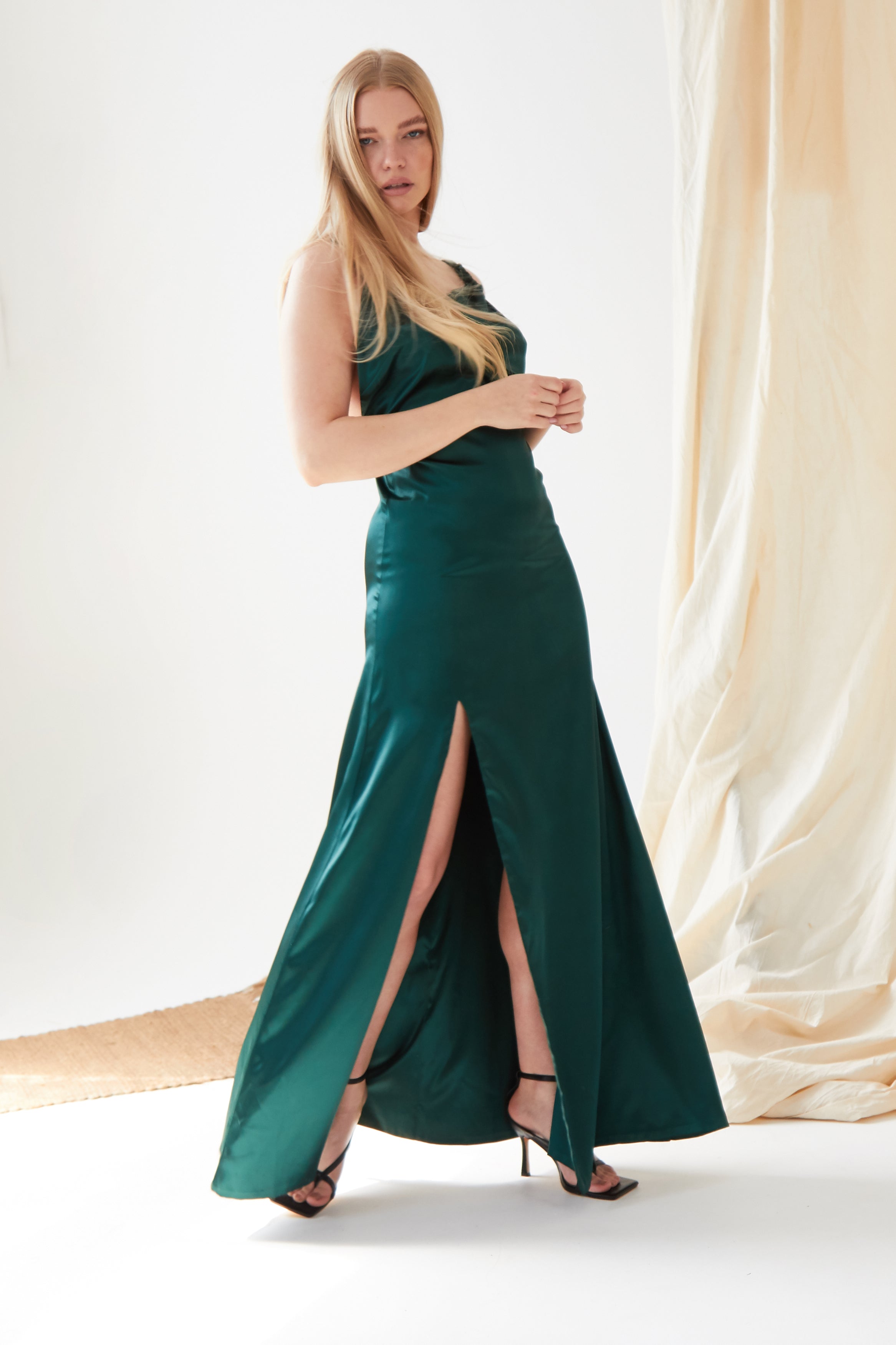 A woman stands indoors in the Sarvin Emerald Green Maxi Dress, featuring high side slits and paired with black high-heeled sandals. Light fabric drapes in the background.