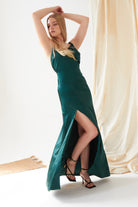A woman poses with arms raised in the Sarvin Emerald Green Maxi Dress, featuring a thigh-high slit. She stands on a woven mat before cream drapes, embodying effortless black-tie elegance.