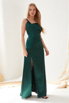 A woman in the Sarvin Emerald Green Maxi Dress stands indoors; its satin fabric and thigh-high slit exude elegance. Her long blonde hair and black open-toe heels complete the chic look against a softly lit, minimal background.
