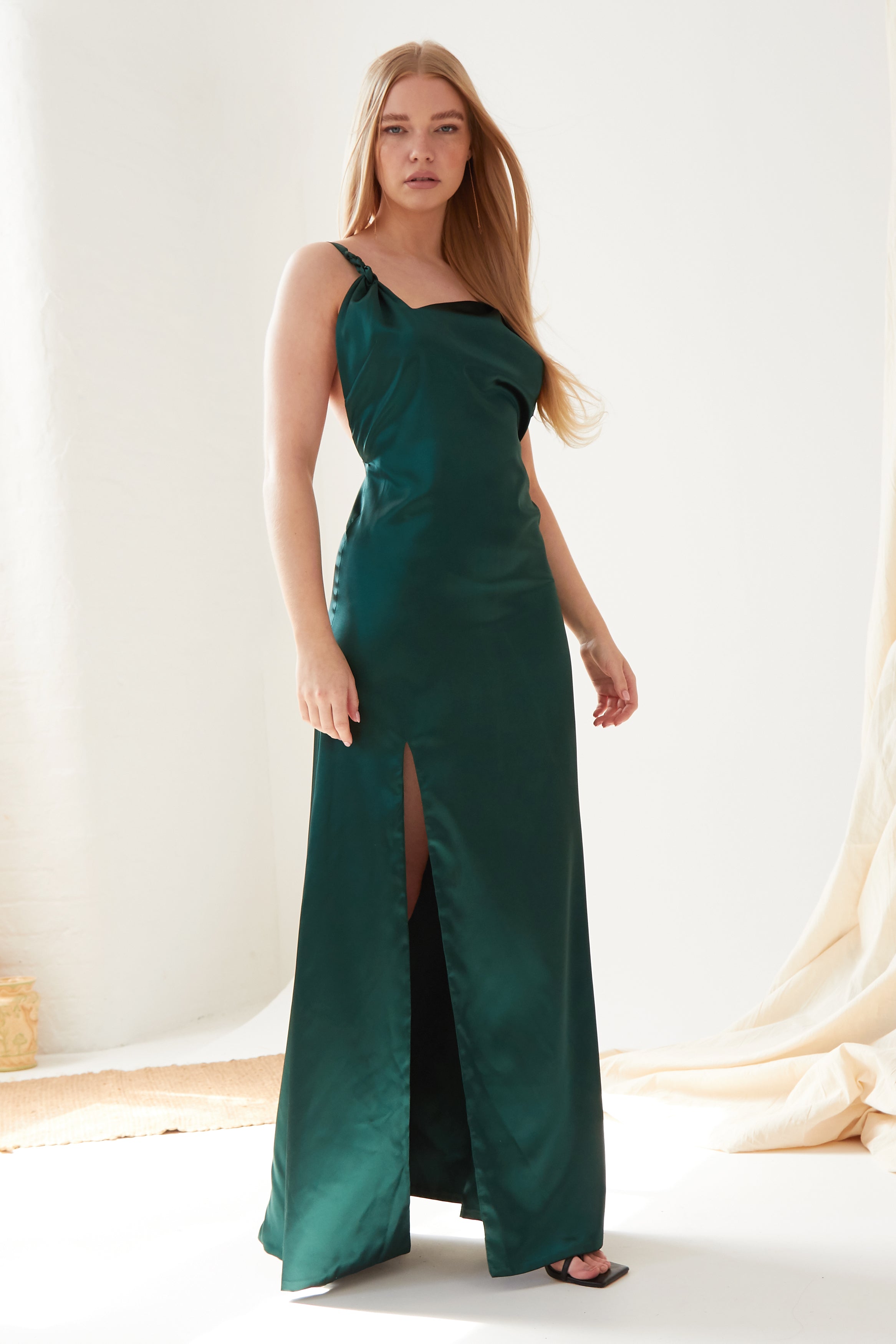 A woman in the Sarvin Emerald Green Maxi Dress stands indoors; its satin fabric and thigh-high slit exude elegance. Her long blonde hair and black open-toe heels complete the chic look against a softly lit, minimal background.