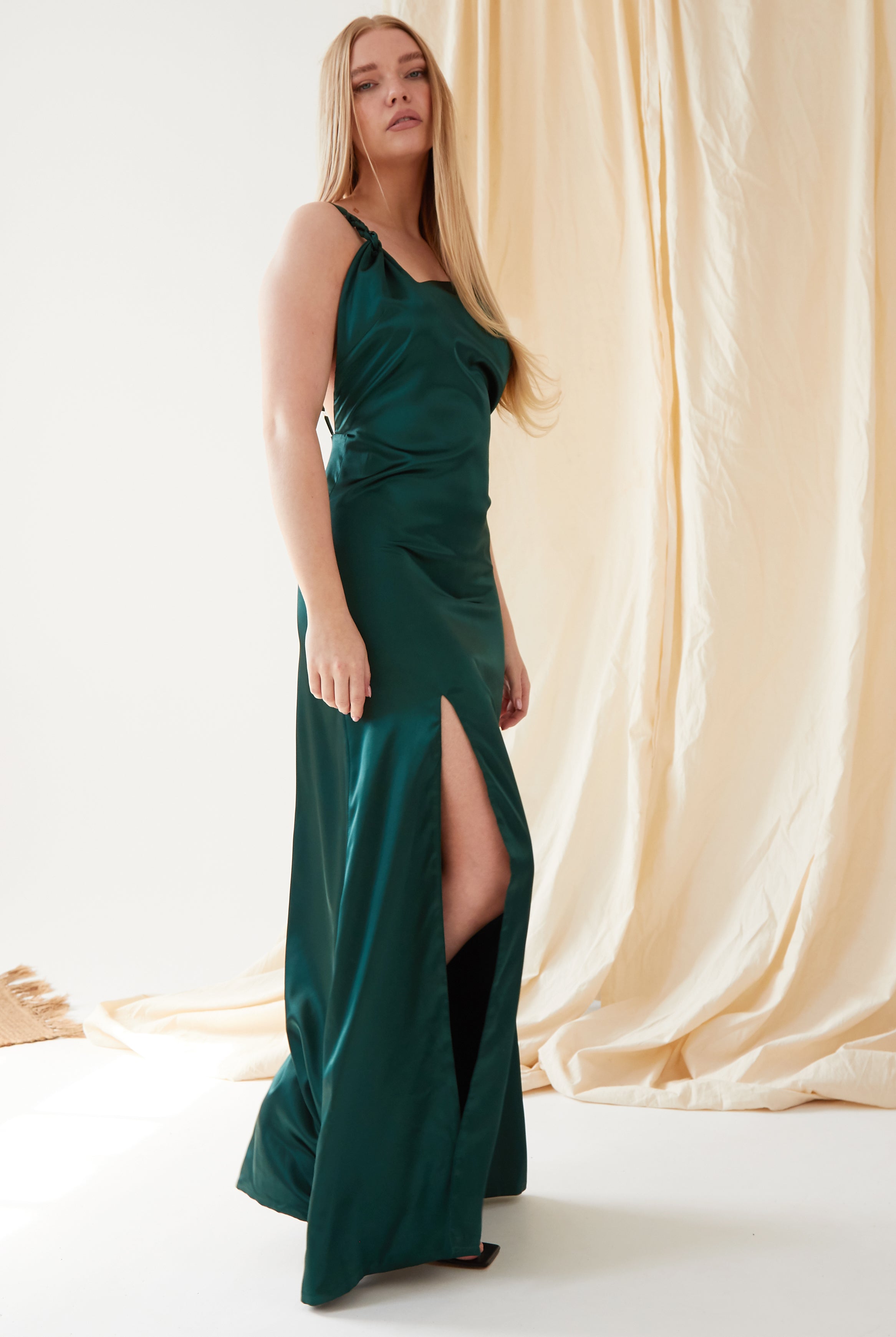 A woman wearing the Sarvin Emerald Green Maxi Dress with a thigh-high slit stands indoors on a white floor, beige drapes behind her. She has long straight blonde hair and black shoes, creating an elegant black-tie look.
