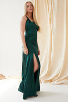 A woman wearing the Sarvin Emerald Green Maxi Dress with a thigh-high slit stands indoors on a white floor, beige drapes behind her. She has long straight blonde hair and black shoes, creating an elegant black-tie look.