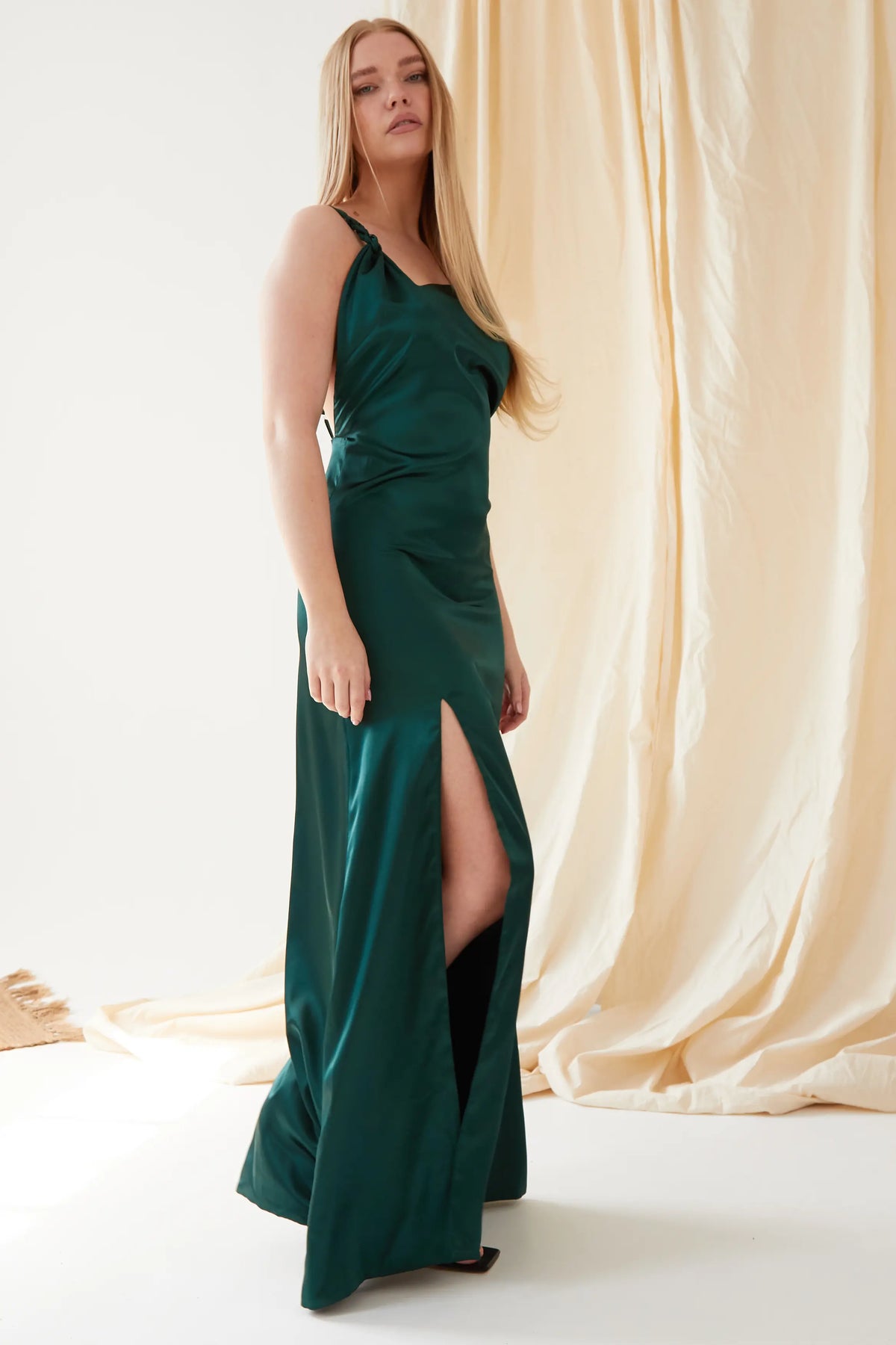 A woman wearing the Sarvin Emerald Green Maxi Dress with a thigh-high slit stands indoors on a white floor, beige drapes behind her. She has long straight blonde hair and black shoes, creating an elegant black-tie look.