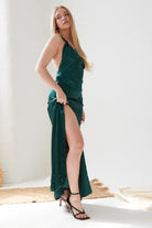 A woman with long blonde hair poses in a Sarvin Emerald Green Maxi Dress, lifting one side to reveal her leg and wearing black heeled sandals in a bright, minimal room.