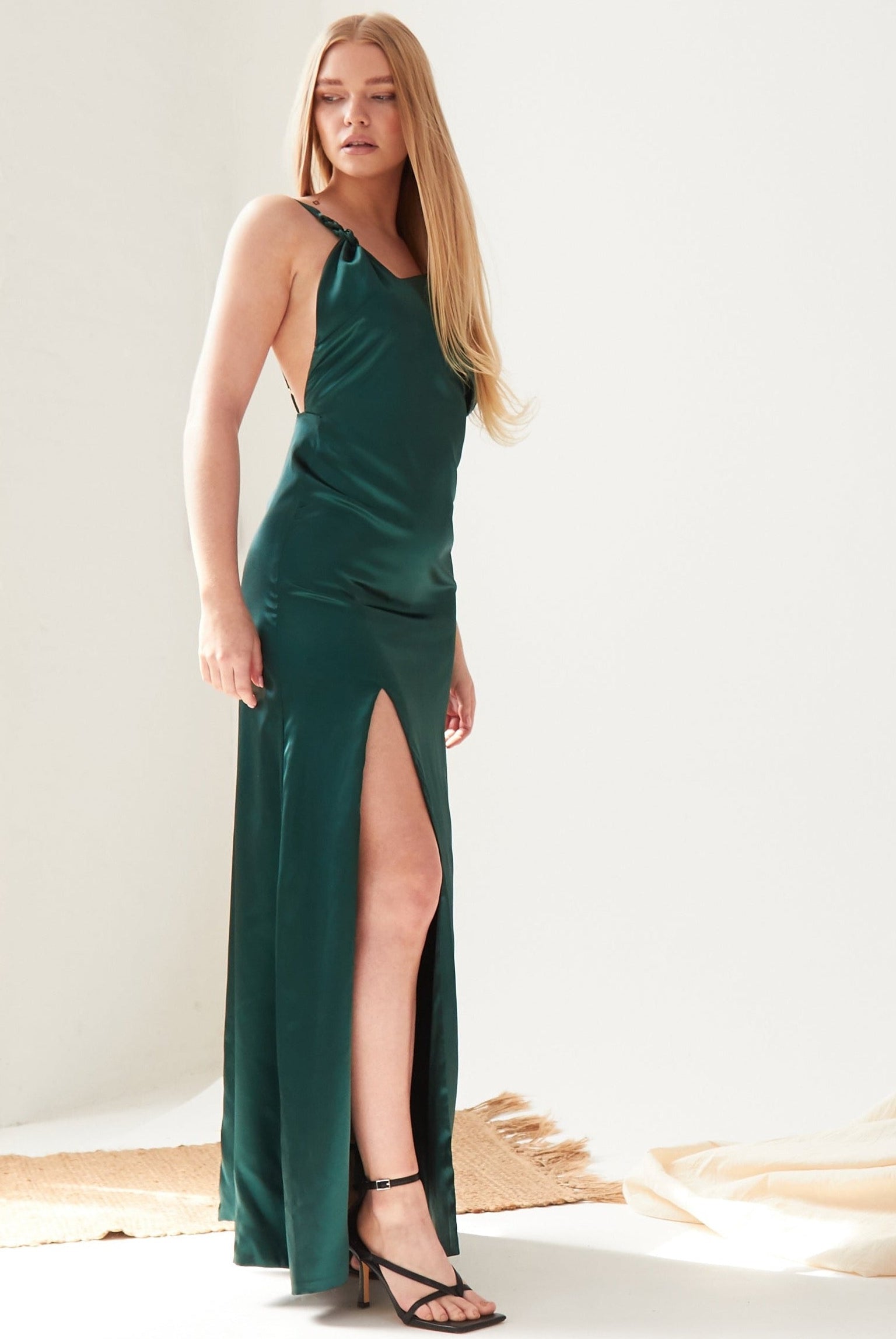 A woman poses indoors against a white background in the Sarvin Emerald Green Maxi Dress, featuring a thigh-high slit and paired with black strappy high-heeled sandals. Her long straight blonde hair complements the minimal decor.