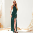 A woman poses indoors against a white background in the Sarvin Emerald Green Maxi Dress, featuring a thigh-high slit and paired with black strappy high-heeled sandals. Her long straight blonde hair complements the minimal decor.