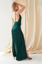 A person with long blond hair models the Sarvin Emerald Green Maxi Dress, featuring a sleeveless design, low back, and high slit, standing in profile before cream-colored draped fabric.