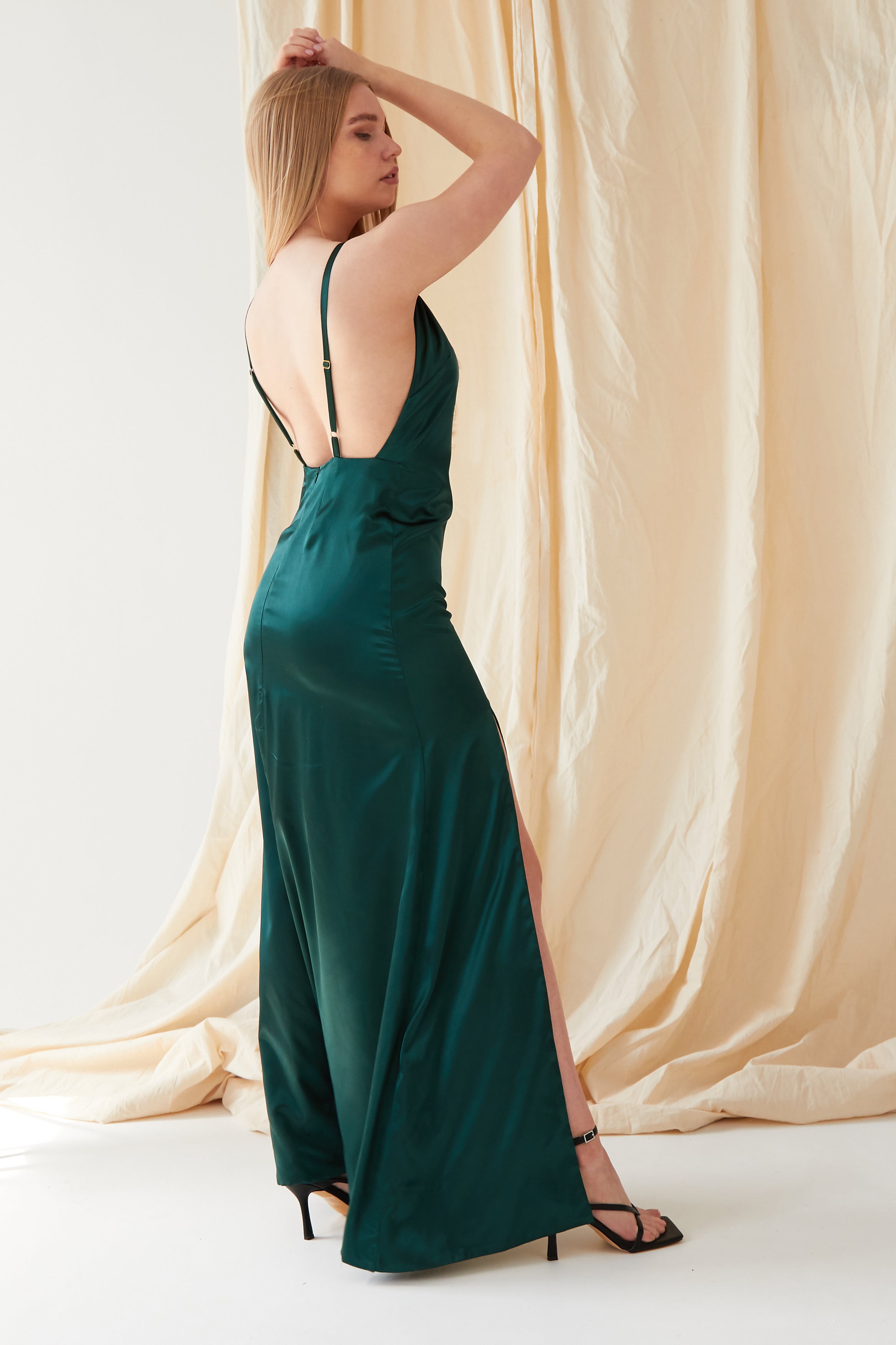 A person with long blond hair models the Sarvin Emerald Green Maxi Dress, featuring a sleeveless design, low back, and high slit, standing in profile before cream-colored draped fabric.