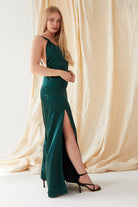 A woman stands before a beige draped backdrop in the Sarvin Emerald Green Maxi Dress, featuring lustrous satin and a high slit. She faces forward with one hand at her waist, radiating classic black-tie elegance.