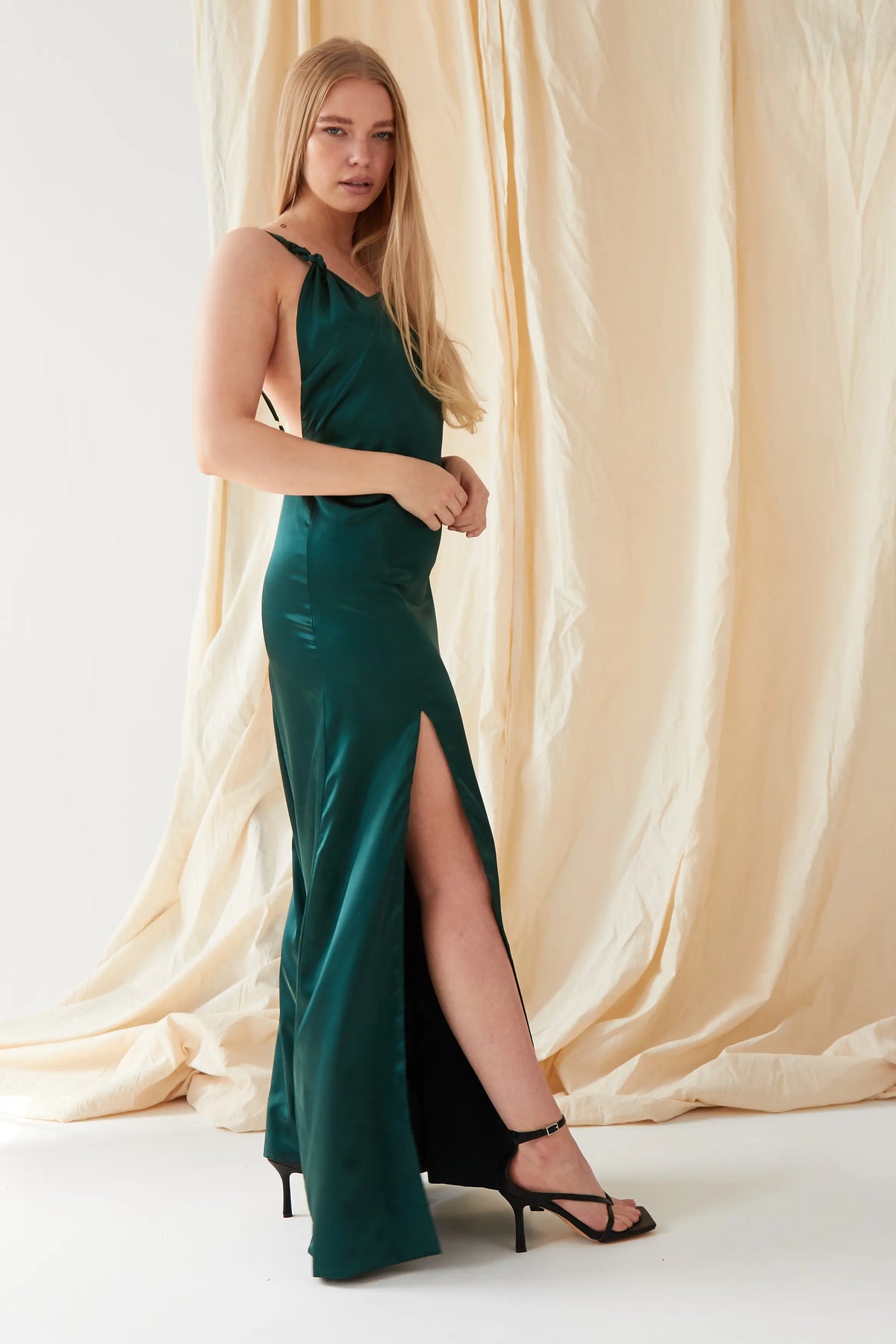 A woman stands before a beige draped backdrop in the Sarvin Emerald Green Maxi Dress, featuring lustrous satin and a high slit. She faces forward with one hand at her waist, radiating classic black-tie elegance.
