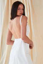 A woman with long dark hair wears the Sarvin A-Line Mini Slip Dress in ivory white, standing before a light draped fabric backdrop and glancing over her shoulder toward the camera.