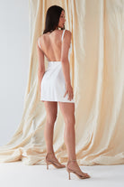 In front of cream fabric, a person wears the Sarvin A-Line Mini Slip Dress in ivory white with nude heels. Facing away with a slight head turn, they showcase the dress's elegant silhouette and low back.