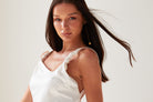 A woman with long brown hair wears a white satin top with ruched straps and Sarvin’s Flower Shape 18K Gold Drop Earrings. Her hair is swept to the side, and she poses against a plain light background with a neutral expression.