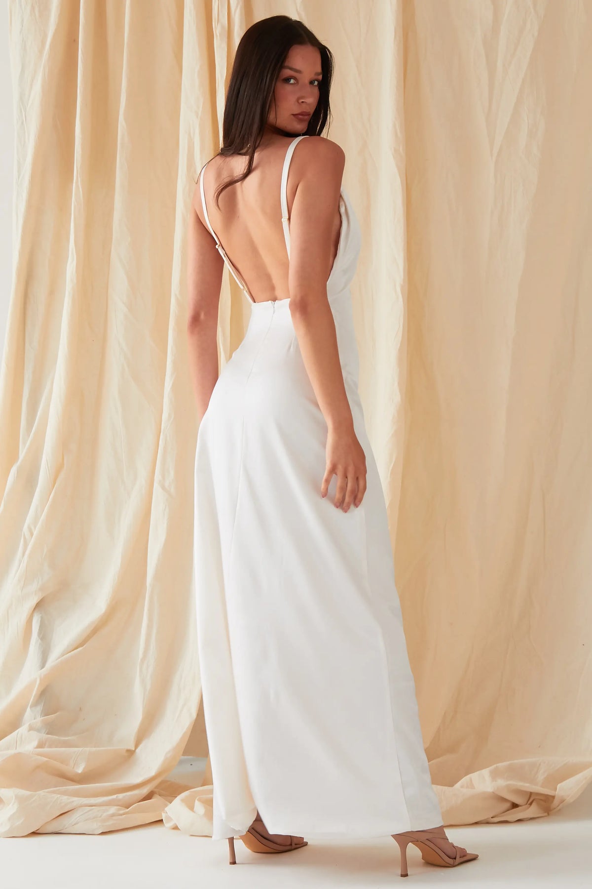 A person with long dark hair wears the Sarvin Ivory Satin Asymmetric Backless Maxi Dress, posing before beige fabric drapes in nude heeled sandals and looking over their shoulder in this elegant evening gown.