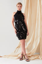 A person stands before beige fabric wearing the Sarvin Sparkly Black Mini Dress with a high neck and strappy black heels. Their light hair is pulled back as they look directly at the camera.