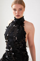 A person with straight, light brown hair models the Sarvin Sparkly Black Mini Dress, featuring large black sequins and a sleeveless design, while looking at the camera against a plain light background.