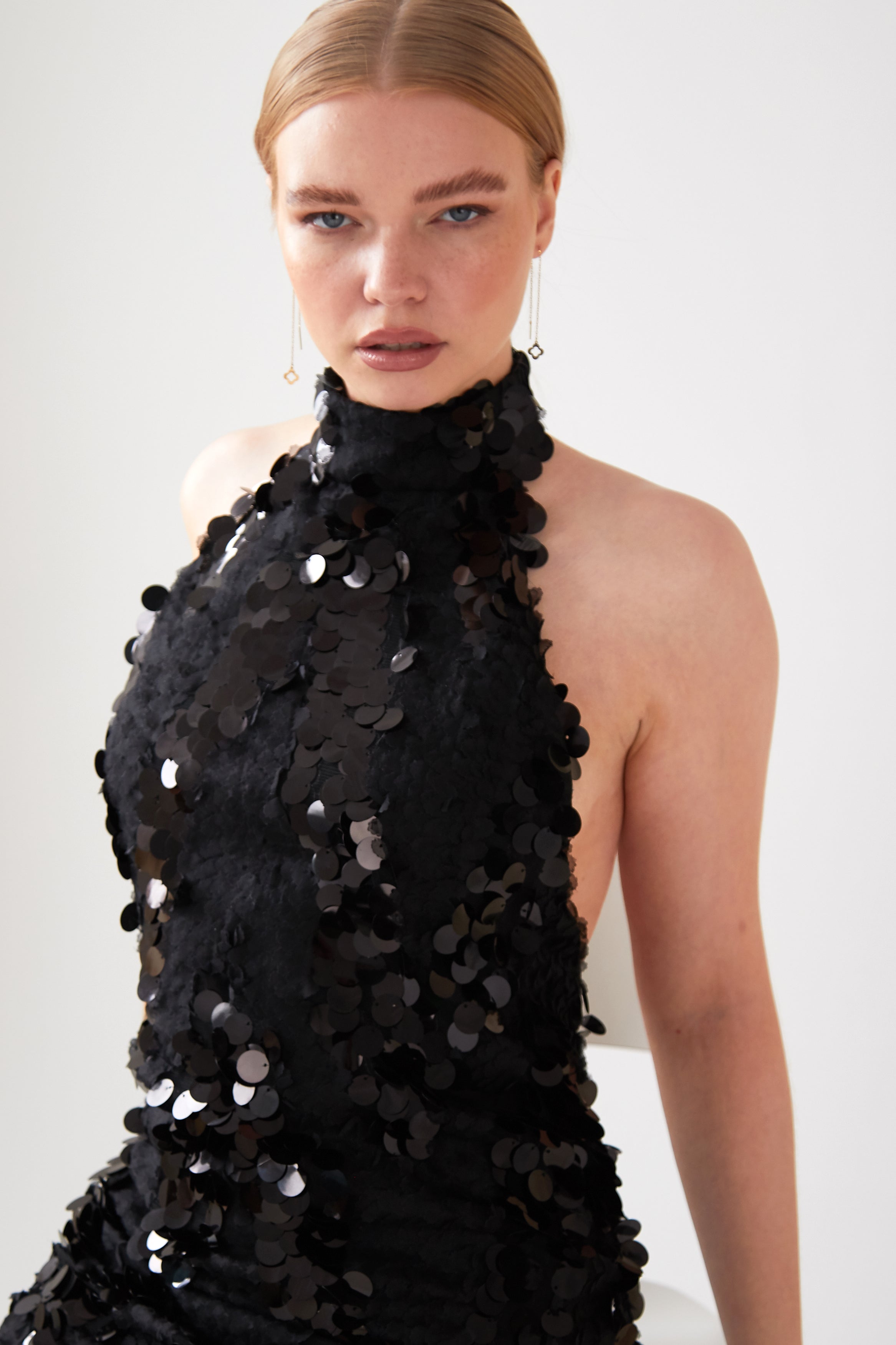 A person with straight, light brown hair models the Sarvin Sparkly Black Mini Dress, featuring large black sequins and a sleeveless design, while looking at the camera against a plain light background.