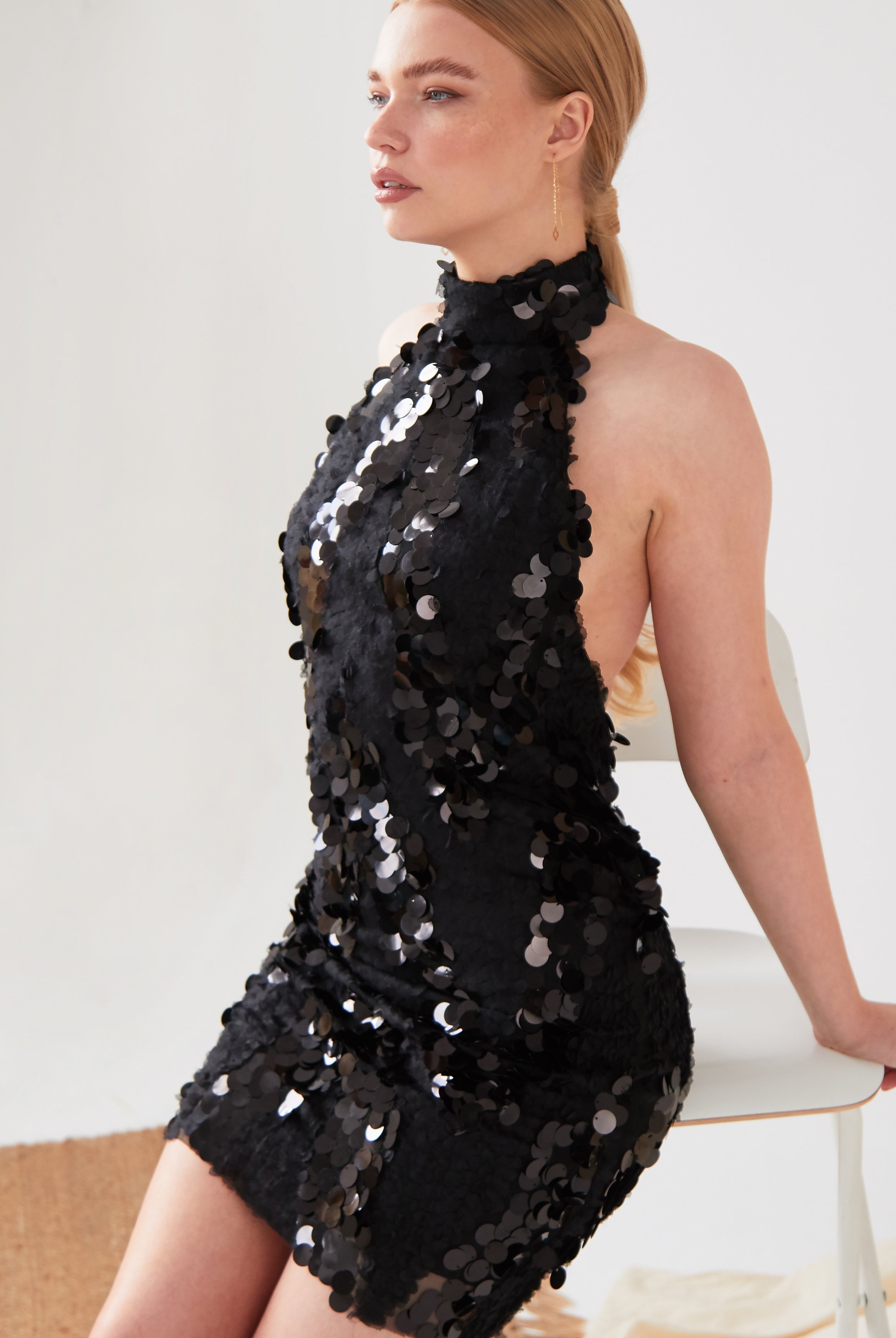 A woman with long blonde hair sits sideways on a white chair, wearing the Sarvin Sparkly Black Mini Dress. She has a neutral expression and looks to the side against a plain, light-colored background.