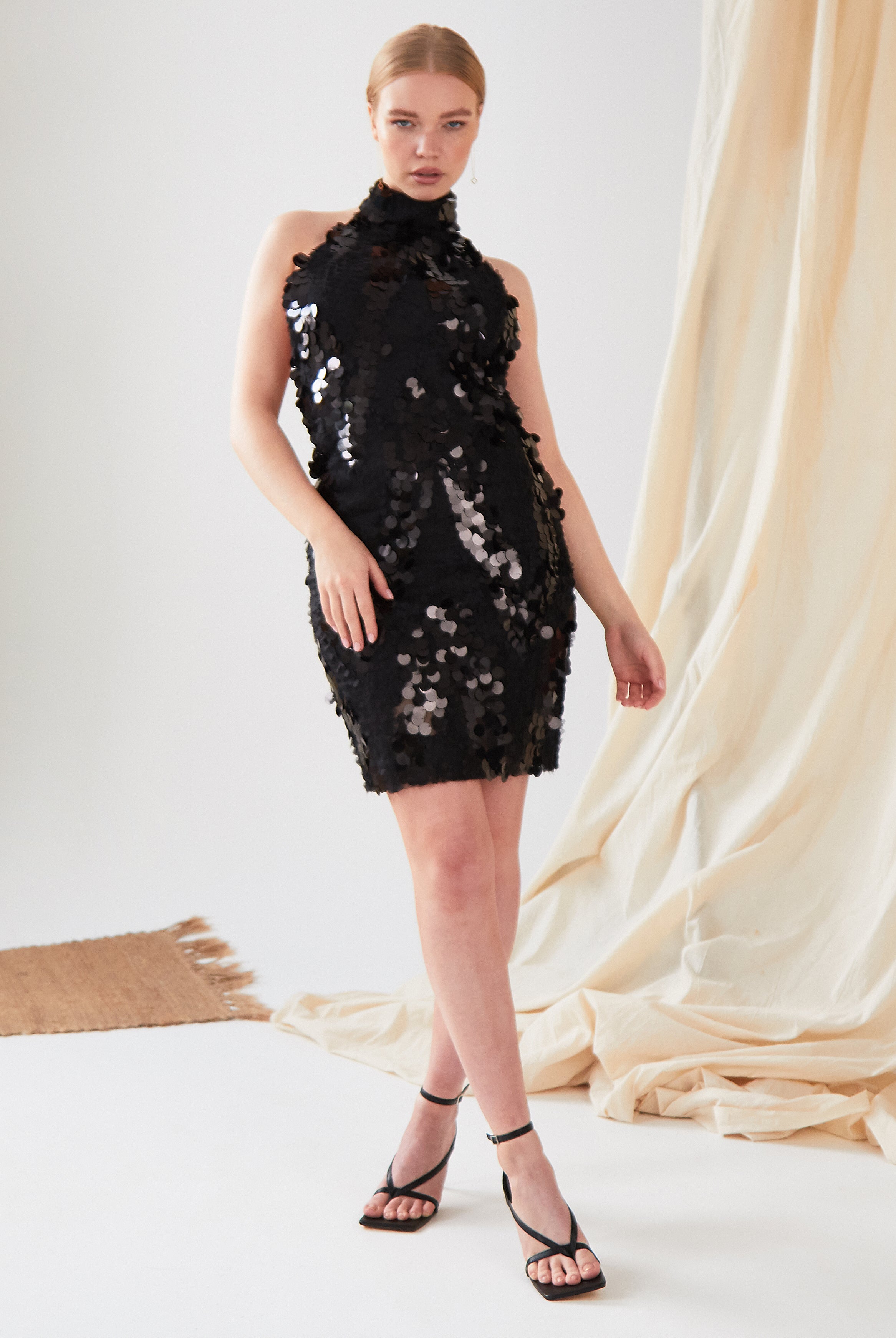 A person stands in a studio wearing the Sarvin Sparkly Black Mini Dress with black high-heeled sandals. A beige fabric backdrop and woven mat add texture to the scene.