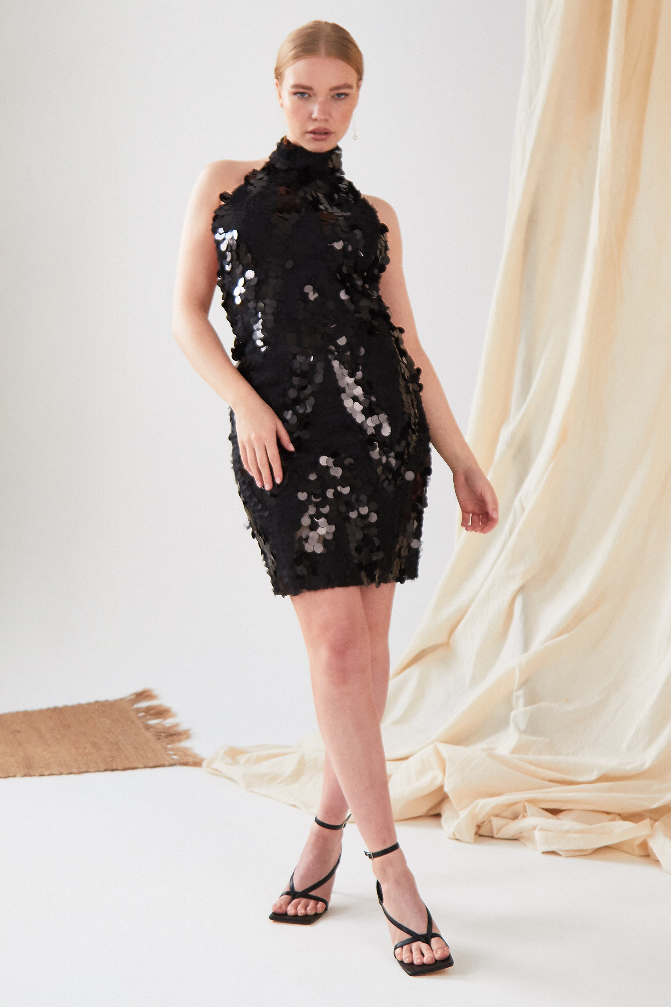 A person stands in a studio wearing the Sarvin Sparkly Black Mini Dress with black high-heeled sandals. A beige fabric backdrop and woven mat add texture to the scene.