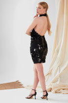 A woman with long blonde hair wears the Sarvin Sparkly Black Mini Dress, an elegant sequined, backless halter mini dress, paired with black high-heeled sandals. She stands on a neutral backdrop with beige draped fabric and a woven mat.
