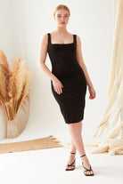A woman stands indoors wearing the Sarvin Black Polka Dot Dress with her hair tied back and black heels, posing with one hand by her side against pampas grass and neutral drapery.