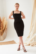 A person poses in a studio wearing the Sarvin Black Polka Dot Dress styled with black heeled sandals. Neutral fabric drapes and pampas grass decor add an elegant touch, making this dress perfect for a wedding guest look.