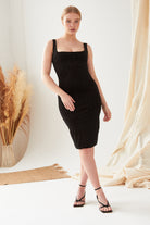 A woman is indoors wearing the Sarvin Black Polka Dot Dress, featuring fitted black velvet with small gold dots, paired with black strappy heels. Neutral pampas grass and beige fabric drapes decorate the background.