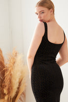A person wears the Sarvin Black Polka Dot Dress, a fitted, sleeveless velvet design. Indoors, they stand turned to the side, looking over their shoulder with tall dried plants in the background.