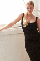 A person with long blonde hair in a ponytail poses indoors by a white window ledge, wearing the Sarvin Black Polka Dot Dress—a fitted, square-neck dress with gold dots, making it an elegant choice for a wedding guest.