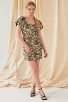A woman stands before a cream fabric backdrop wearing the Sarvin Puff Shoulder Mini Dress, featuring green and white hues, one sleeve, and puff shoulders. She styles her long blonde hair in a ponytail with black strappy heels.