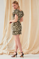 A woman poses before beige draped fabric, glancing over her shoulder in the Sarvin Puff Shoulder Mini Dress—golden floral jacquard—paired with black high-heeled sandals.