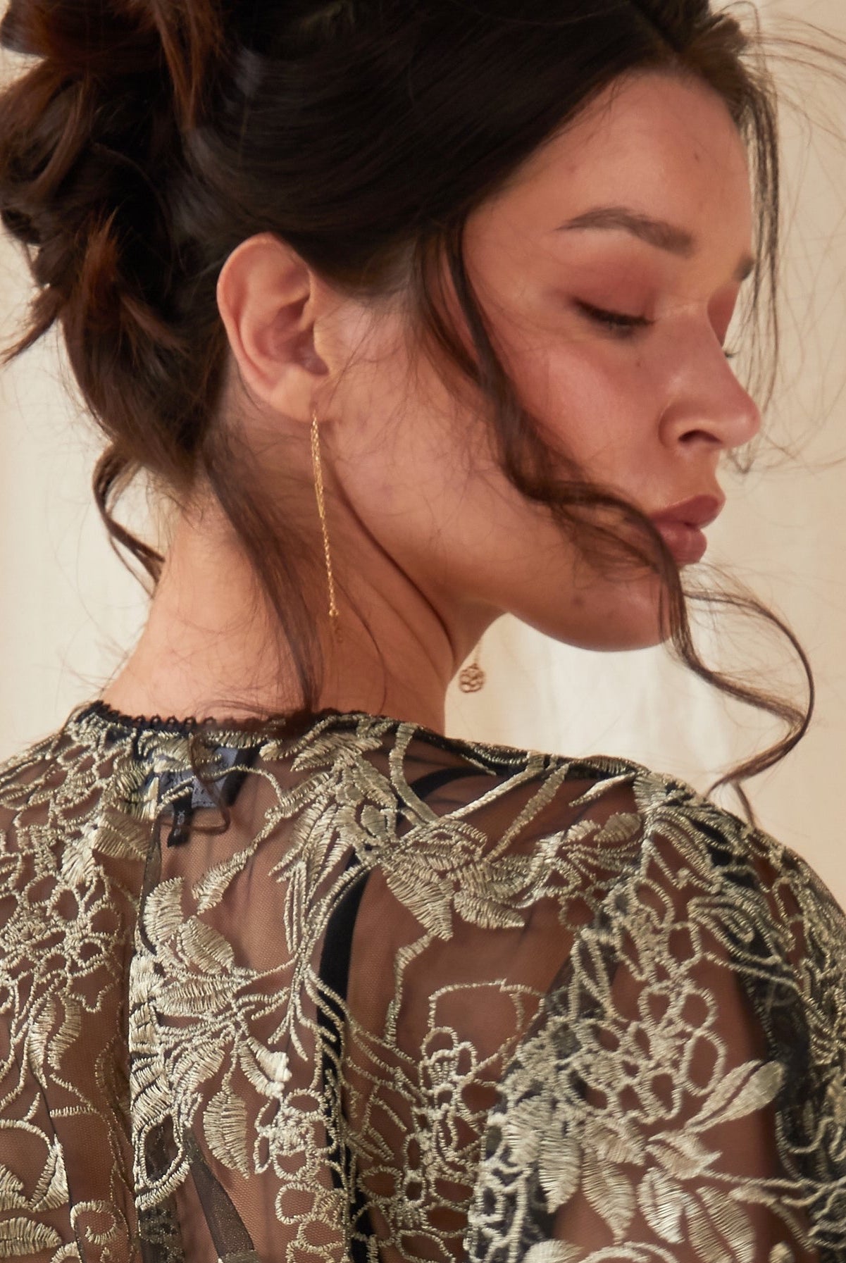 A woman with dark hair in an updo faces away, eyes closed, wearing a sheer black top with gold embroidery. She showcases Sarvin's Flower Shape 18K Gold Drop Earrings against a softly lit neutral background.