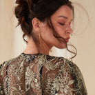 A woman with dark hair in an updo faces away, eyes closed, wearing a sheer black top with gold embroidery. She showcases Sarvin's Flower Shape 18K Gold Drop Earrings against a softly lit neutral background.